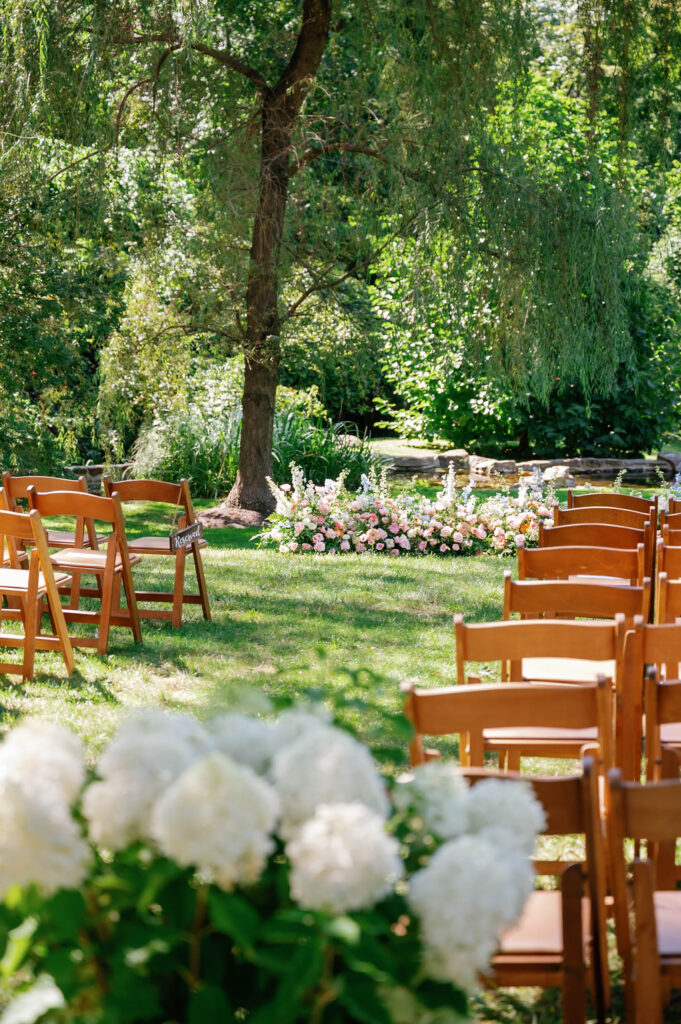 Outdoor garden ceremony setup at Appleford Estate in Villanova, Pennsylvania with wooden chairs arranged beneath towering trees and soft floral arrangements lining the aisle.