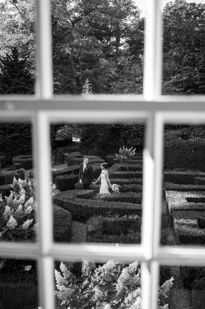 Black and white portrait of the couple walking through the boxwood garden paths at Appleford Estate, photographed through the manor house window.