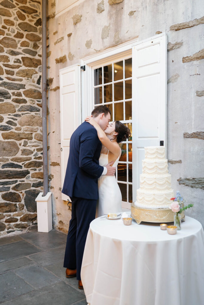 Bride and groom sharing a kiss beside their wedding cake outside the historic stone walls of Appleford Estate.